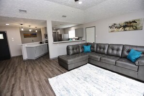 A modern living room with a sectional sofa, a kitchen area, and a dining table. - Modern Spacious Home in Quiet Neighborhood (La Porte)