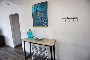 Una habitación con una mesa de madera, un cuadro de pavo real, un cartel de bienvenida y un perchero metálico.