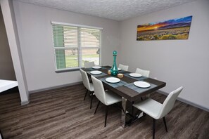 Un comedor con una mesa dispuesta para cuatro personas, sillas blancas, un florero turquesa y un cuadro con un atardecer en la pared.
