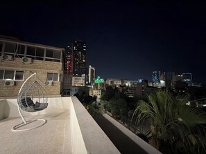 Una terraza en la azotea con una silla colgante, vista del horizonte urbano nocturno y un edificio con un letrero verde.