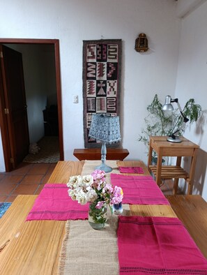 A dining area with a wooden table, pink placemats, a vase with flowers, a wall hanging, and a lamp.