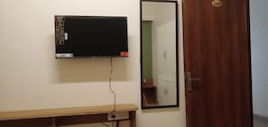 A room with a mounted television, a wooden bench, a mirror, and a door with a number plate. - Laya Crest (Chennai)