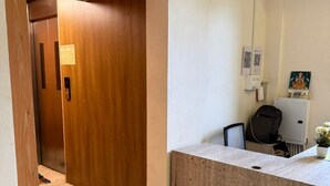 A reception area with a wooden door, a small desk, a chair, a potted plant, and a security camera. - Laya Crest (Chennai)