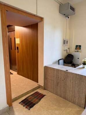 A reception area with a wooden door, a small desk, a chair, a potted plant, and a security camera. - Laya Crest (Chennai)