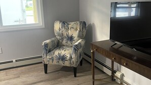 Olohuone, jossa on kukkapainokuosilla verhoiltu nojatuoli, televisio puisen jalustan päällä, kuvioitu matto ja ikkuna, jossa on sälekaihtimet.