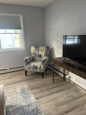 Ein Wohnzimmer mit einem blumigen Sessel, einem Fernseher auf einem Holzständer, einem gemusterten Teppich und einem Fenster mit Jalousien.