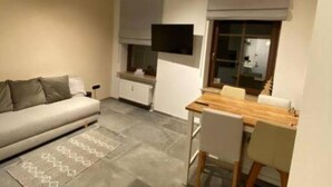 Ett modernt vardagsrum med en soffa, en matplats med bord och stolar, en tv som är monterad på väggen och ett fönster med persienner.