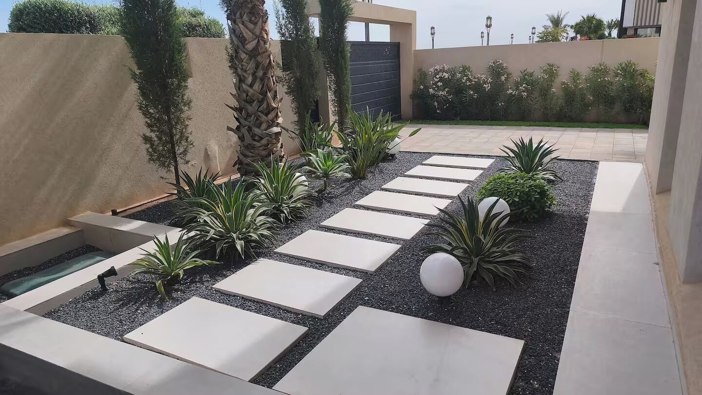 A modern outdoor patio with geometric stone pathways, palm trees, and a variety of shrubs.