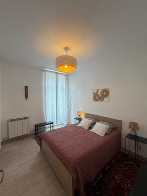 A bedroom with a bed, a radiator, a window with curtains, a bedside table with a lamp, and a wall art piece. - Apartment with breathtaking view of the vineyard and quiet village center (Montréal)