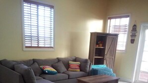 A living room with a grey sectional sofa, a wooden coffee table, and a blue armchair. There is a window with blinds, a door, and a shelf with items. - Anchorage - 4BR Luxury Beach House in Exuma – 50ft to calm water, Sleeps 10 (Black Point)