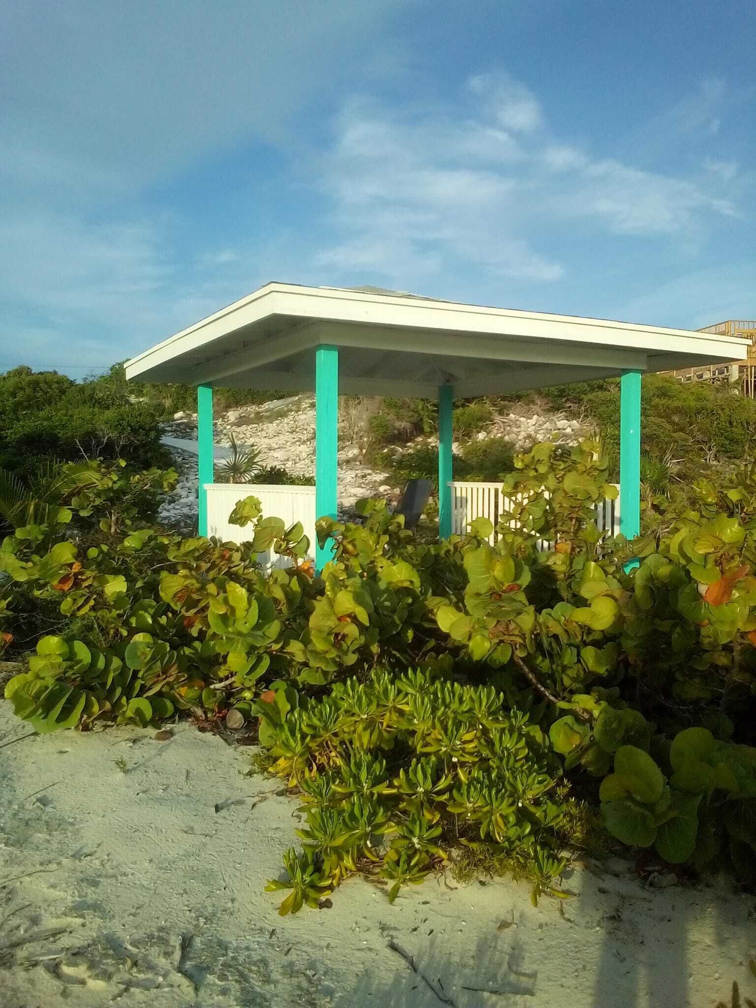 Un espace extérieur abrité, doté d’un toit blanc et de piliers turquoise, entouré de verdure et d’un sol sablonneux.