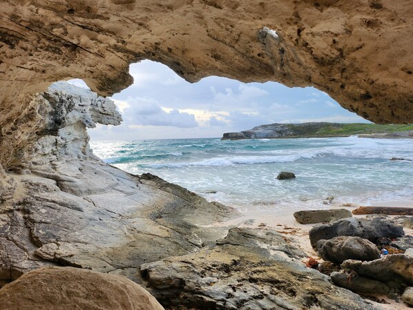 Eine Küstenlandschaft mit einer natürlichen Felsformation, die sich über das Meer erstreckt, einem Sandstrand und einer in der Ferne liegenden Felsklippe.
