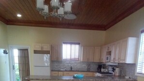 A dining area with a table set for four, a kitchen in the background, and a ceiling fan. - Anchorage - 4BR Luxury Beach House in Exuma – 50ft to calm water, Sleeps 10 (Black Point)