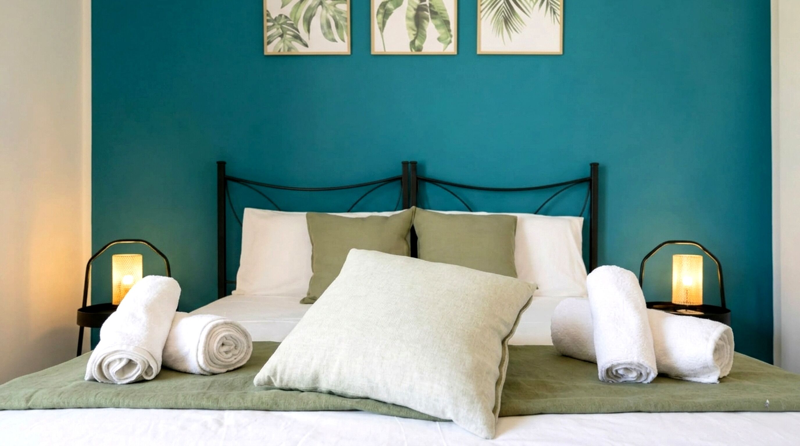 Een bed met kussens en opgerolde handdoeken, een groenblauwe muur en een wanddecoratie van planten.