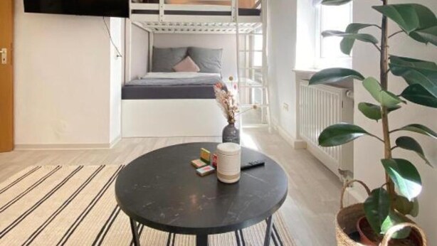 Une chambre moderne avec un lit superposé, une table ronde noire, une plante en pot et un téléviseur fixé au mur.