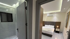 Ein modernes Hotelzimmer mit Duschkabine aus Glas, einem Bett mit Kopfteil, einem Nachttisch und einer wandmontierten Klimaanlage.