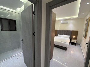 A modern hotel room with a glass shower enclosure, a bed with a headboard, a bedside table, and a wall-mounted air conditioner. - 302 JRE Rooftop With Stunning Boulevard Veiw (Amman)