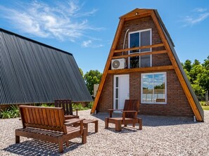 A triangular wooden cabin with a sloped roof, a door, and windows. There are two wooden chairs and a table outside.