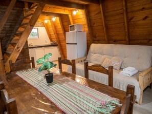 A cozy attic room with a wooden ceiling, a dining table with a woven placemat, a sofa with a blanket, and a white refrigerator.