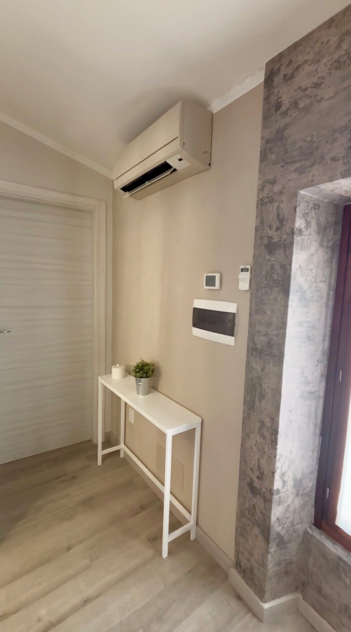 Habitació amb un aparell d'aire condicionat blanc, una taula petita blanca amb una planta, un termòstat fixat a la paret i una porta amb una finestra.