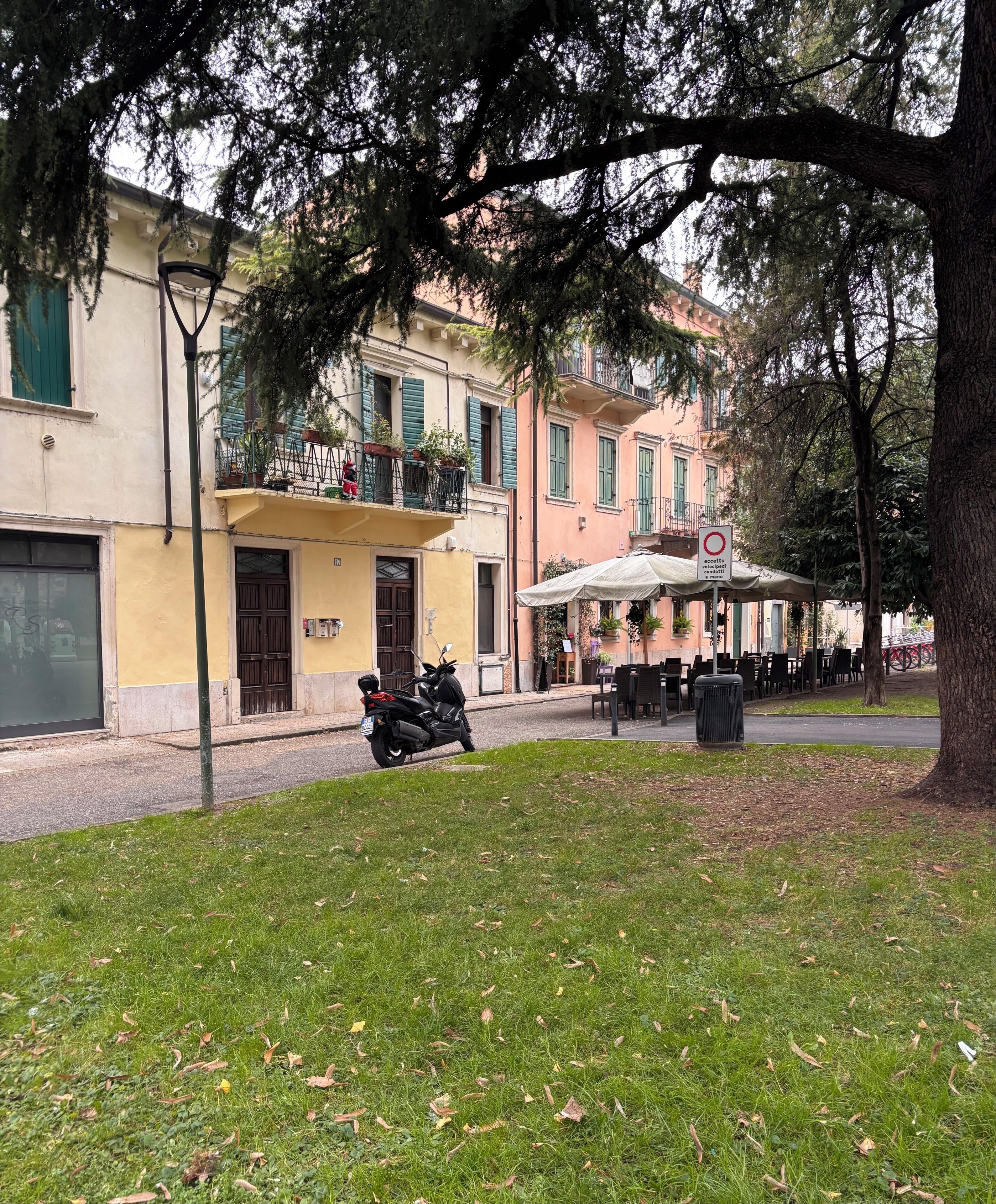 A street with buildings, a parked scooter, and a grassy area with trees.
