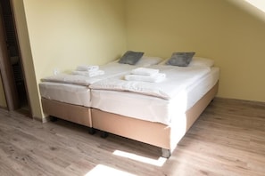 Una habitación con dos camas, cada una con sábanas y almohadas blancas, suelo de madera y pared de tono claro.
