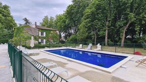 Una piscina rectangular amb una tanca verda, seients de bany de sol blancs i una casa al fons.