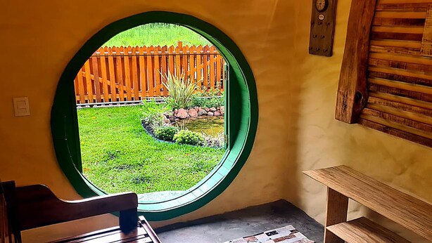 A room with a circular window, a wooden bench, and a wall-mounted clock. - CABANA TEMÁTICA DO HOBBIT - GRAMADO RURAL (Gramado)