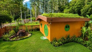 A small, single-story building with a flat roof, surrounded by lush greenery and a wooden fence. - CABANA TEMÁTICA DO HOBBIT - GRAMADO RURAL (Gramado)