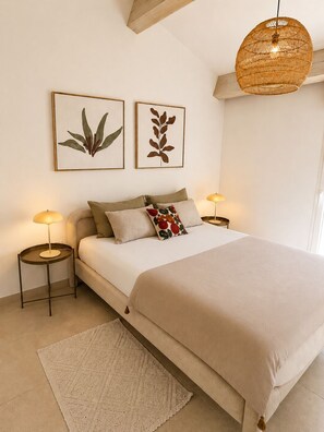 Une chambre à coucher comprenant un lit, deux tables de chevet, un luminaire suspendu et deux reproductions encadrées de motifs botaniques accrochées au mur.