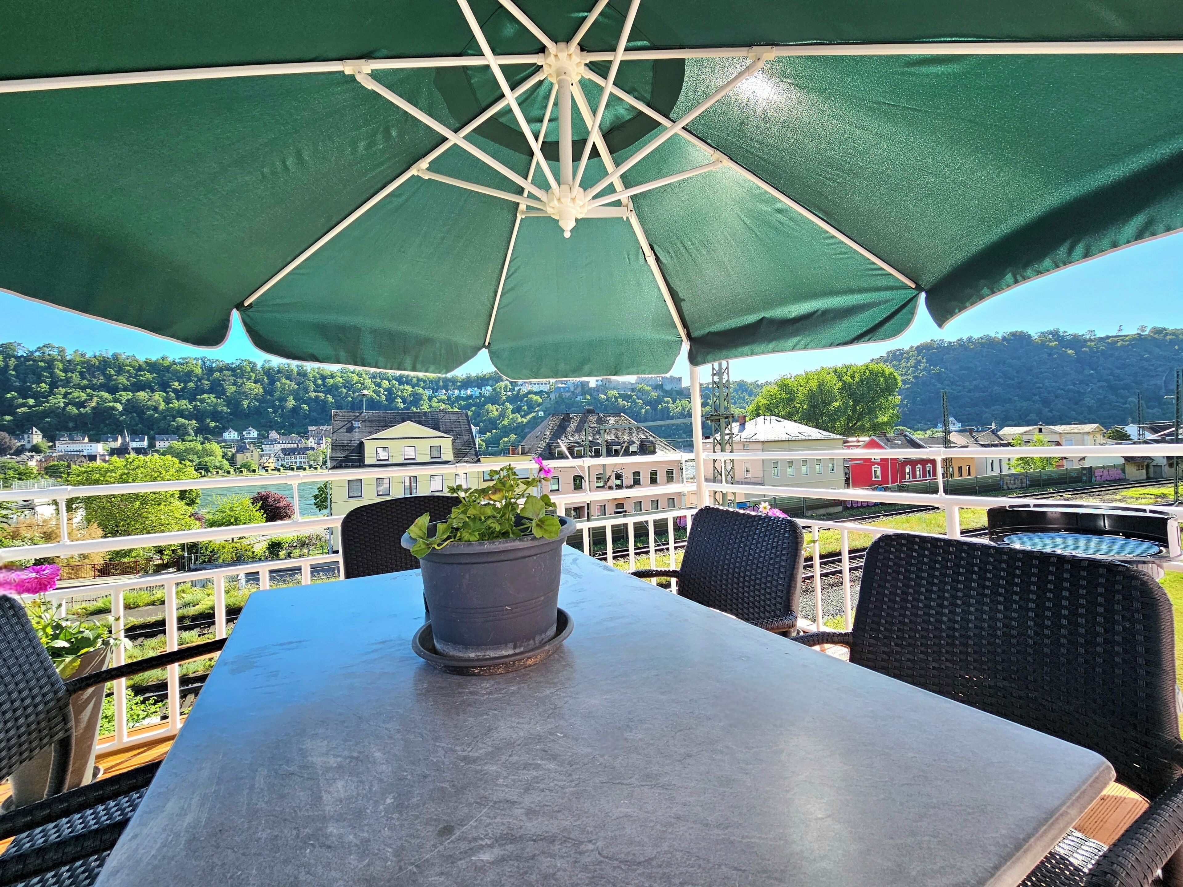 An outdoor seating area with a table and chairs under a green umbrella, overlooking a scenic view of buildings and greenery.