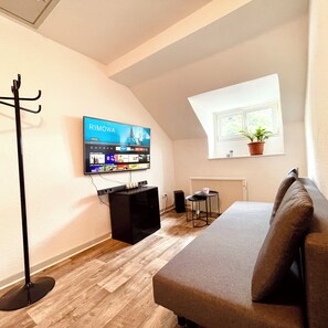 A modern living room with a flat-screen TV, a sofa, a coffee table, a floor lamp, and a potted plant by a window.