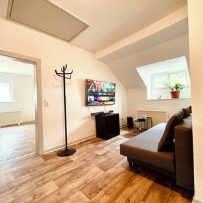 A living room with a flat-screen TV, a coat rack, a sofa, a small table, and a window with a plant.