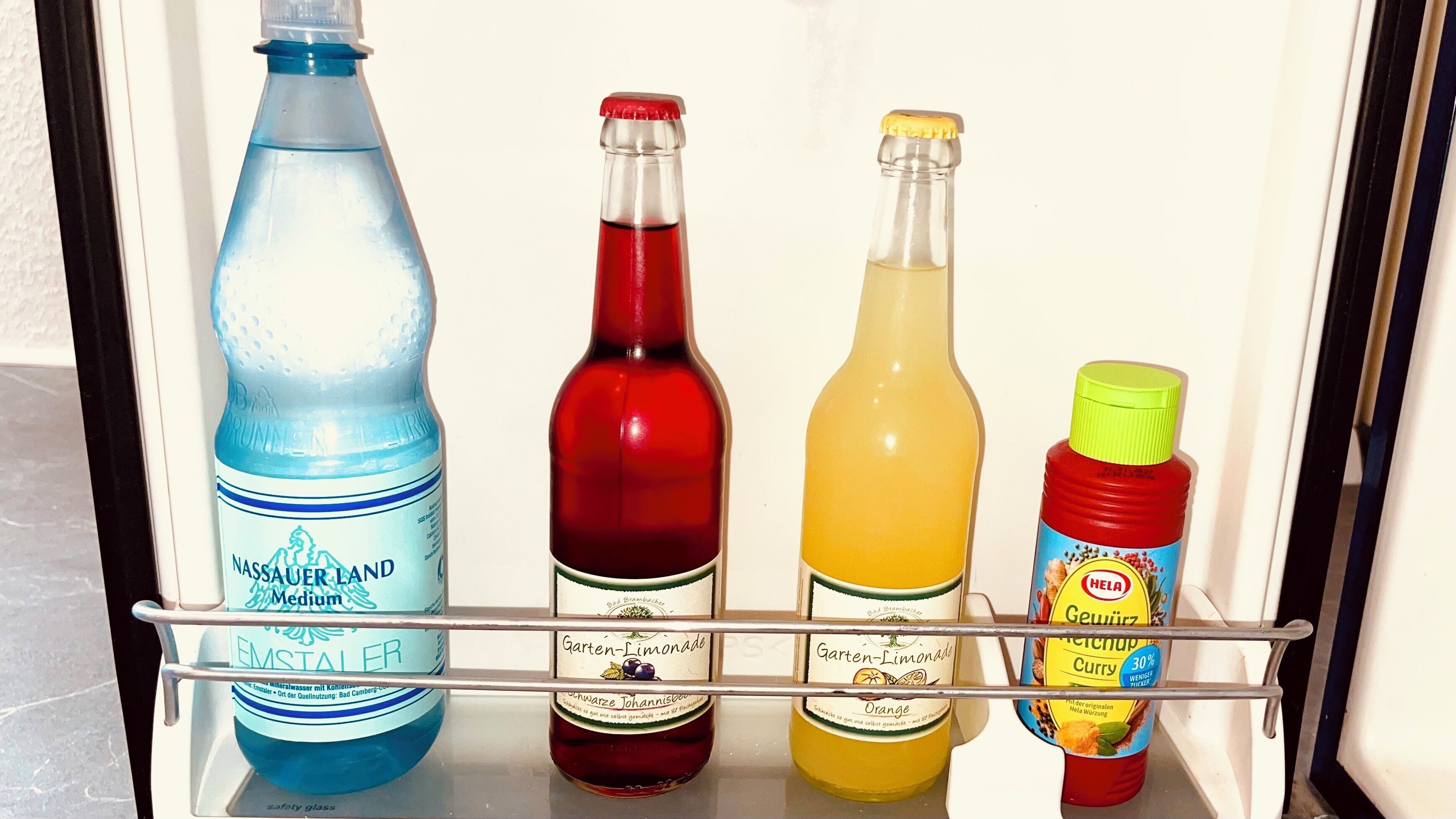 A refrigerator shelf with four bottles: a blue water bottle, a red wine bottle, a yellow juice bottle, and a red ketchup bottle.