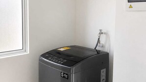 Una lavatrice compatta e moderna in un angolo, con una finestra, una stendibiancheria a parete e un cartello di avvertimento.