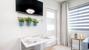 Quarto moderno com televisão de tela plana, uma mesa de jantar branca, plantas em vasos, uma pequena mesa redonda com objetos decorativos e uma janela com persianas.