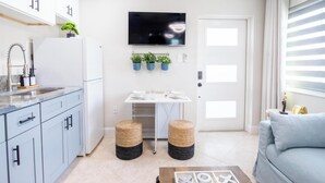 Uma cozinha moderna com televisão integrada, um espaço de jantar com mesa e cadeiras, e uma sala de estar com sofá e janela com persianas.