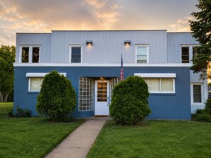 A two-story building with a blue exterior, a front door with a glass panel, and two symmetrical trees in the front yard.