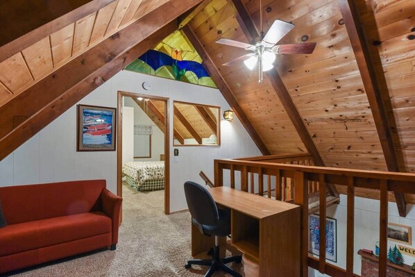 A loft bedroom with a sloped ceiling, a ceiling fan, a desk, a red sofa, and a bed with a checkered bedspread. - Moose Cabin Ideally Located (Cedar Flat)