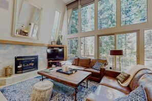 Ein geräumiges Wohnzimmer mit Kamin, Ledersofas, einem Couchtisch und großen Fenstern mit Blick auf einen Wald.