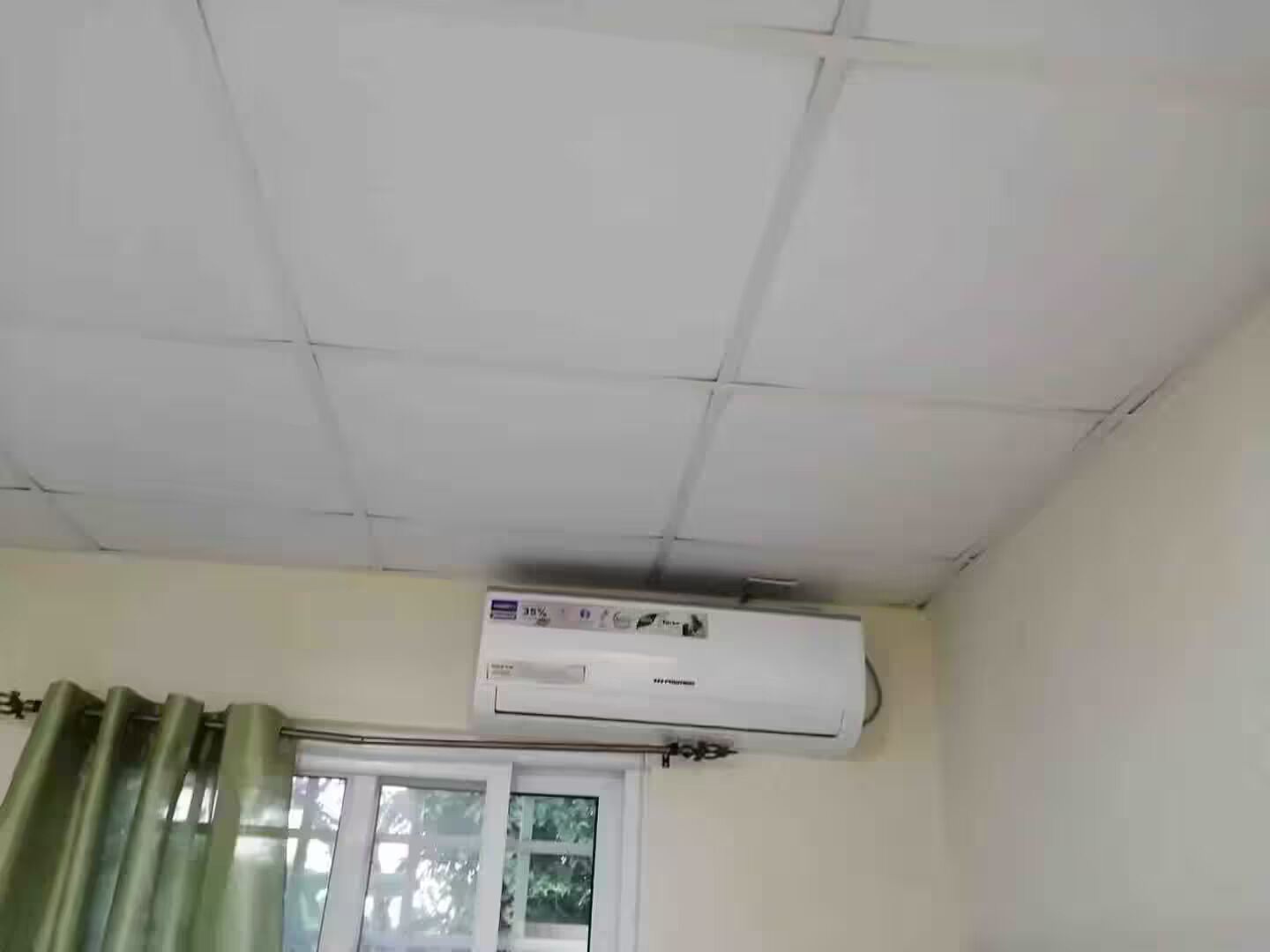 A room with a ceiling-mounted air conditioning unit, a window with curtains, and a wall-mounted thermostat.