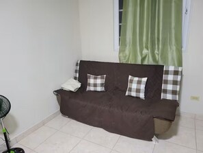 A brown sofa with plaid cushions, a standing fan, and a window with green curtains.