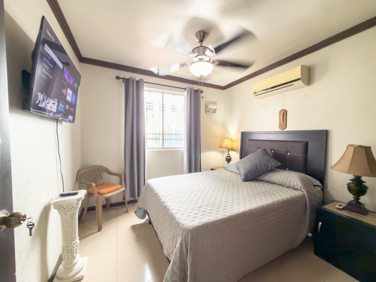 A hotel room with a bed, a ceiling fan, a TV mounted on the wall, a window with curtains, and a nightstand with a lamp.
