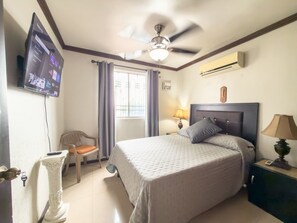 Quarto de hotel com cama, ventoinha no teto, televisão na parede, janela com cortinas e mesa de cabeceira com abajur.