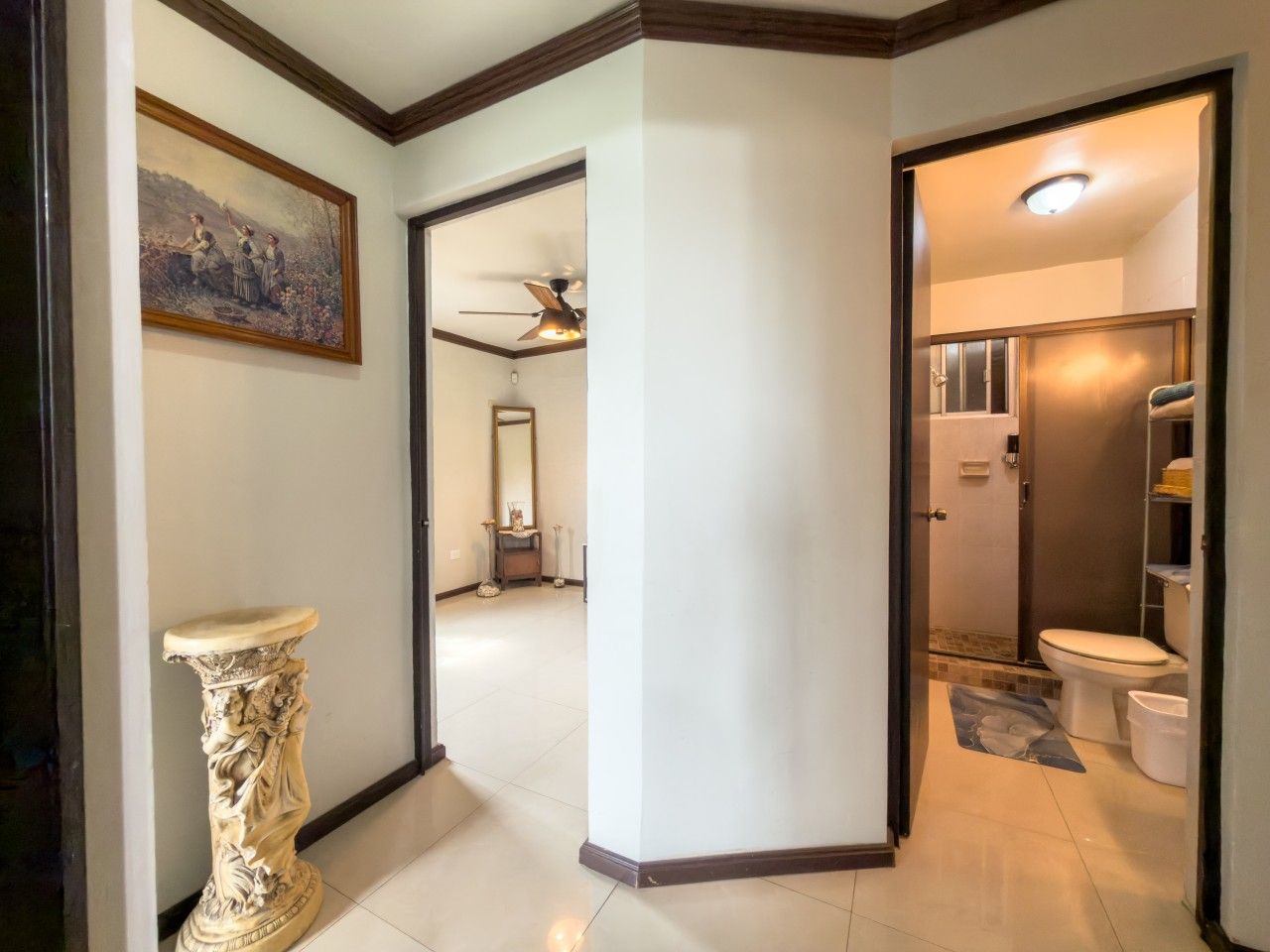 A hallway leading to a bathroom with a toilet, a shower, and a ceiling fan.