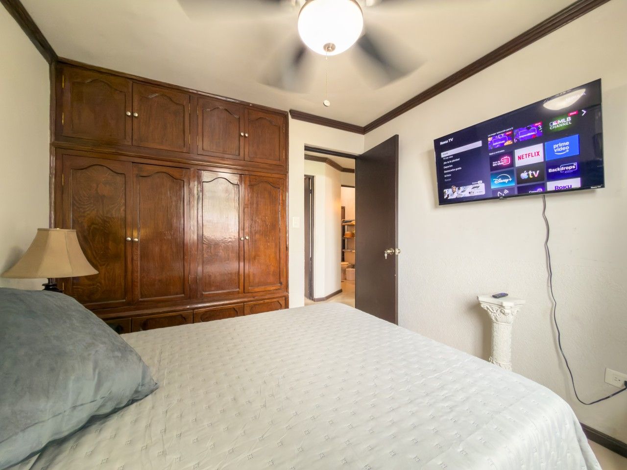 A bedroom with a bed, a wooden wardrobe, a television mounted on the wall, and a white cat statue.