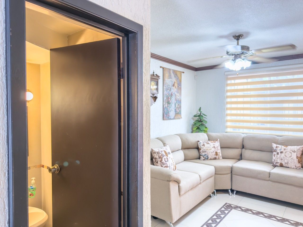 Una sala de estar con un sofá seccional color beige, cojines estampados, un ventilador de techo y una puerta que da a un baño.