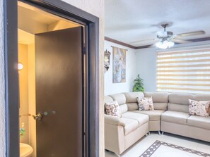 A living room with a beige sectional sofa, patterned cushions, a ceiling fan, and a door leading to a bathroom.