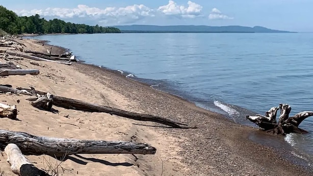 A beach with driftwood, calm water, and distant mountains. - Copper Ridge at Miller's Cottages (Ontonagon)