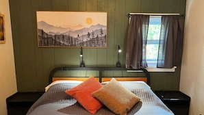 A bedroom with a bed, two nightstands, a painting, and a window with curtains. - Copper Ridge at Miller's Cottages (Ontonagon)
